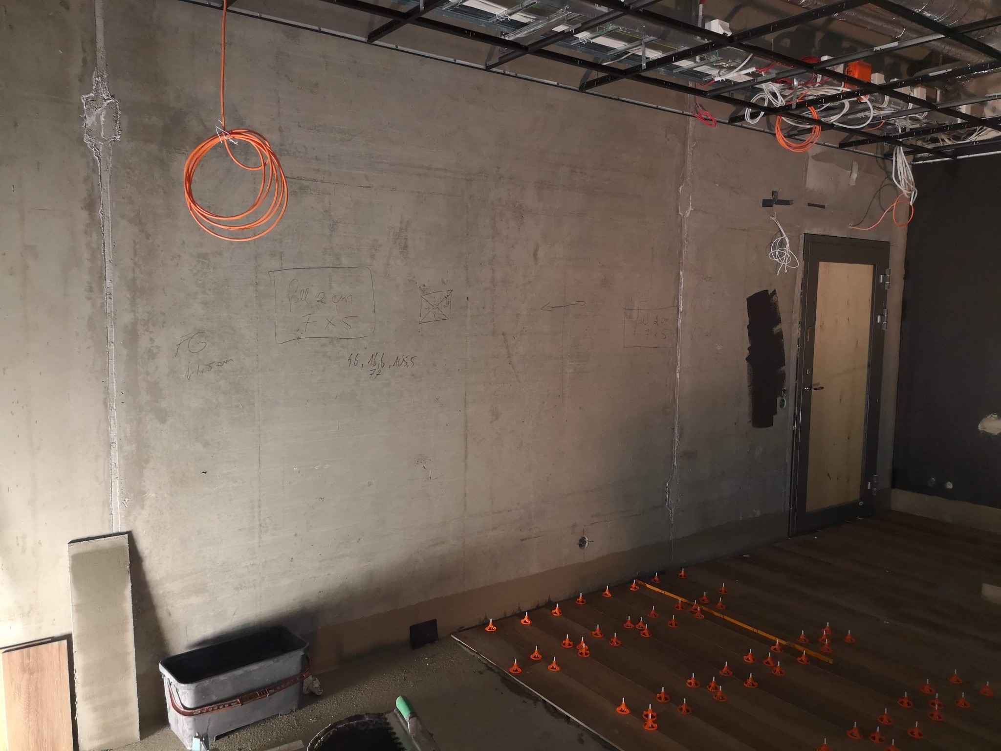 Wnętrze w trakcie układania podłogi z płytek imitujących drewno, widoczne pomarańczowe kliny poziomujące, szare ściany z odręcznymi notatkami, instalacje pod sufitem.