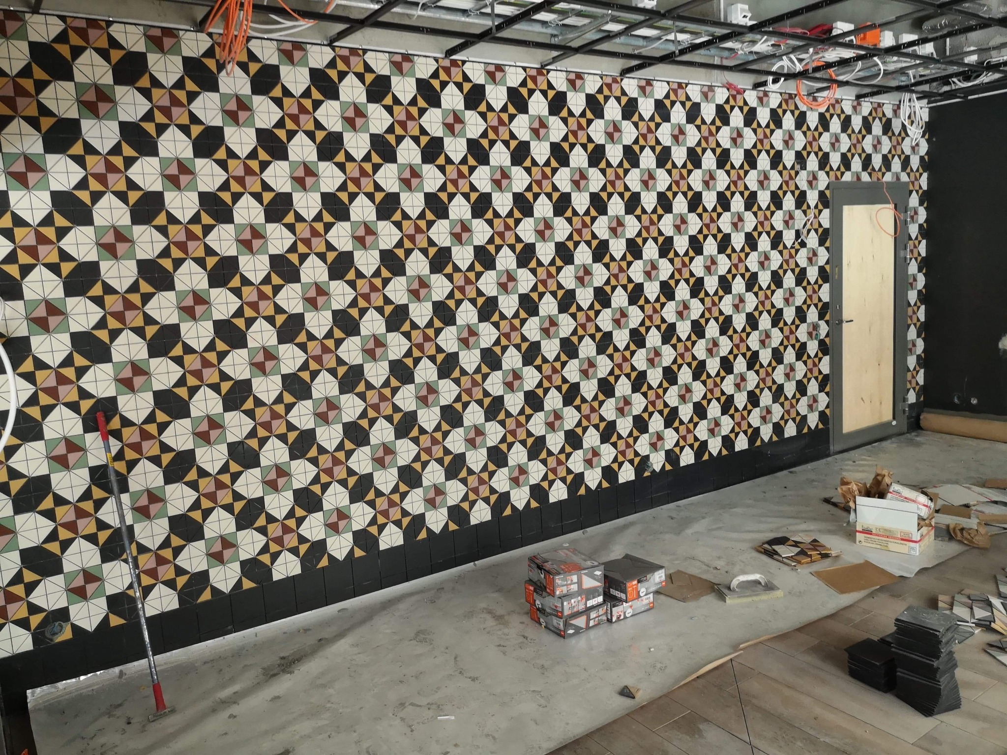 Ściana wyłożona wzorzystymi, kolorowymi płytkami w trakcie montażu, widoczne paczki z płytkami, narzędzia i niedokończone wykończenie pod sufitem.
