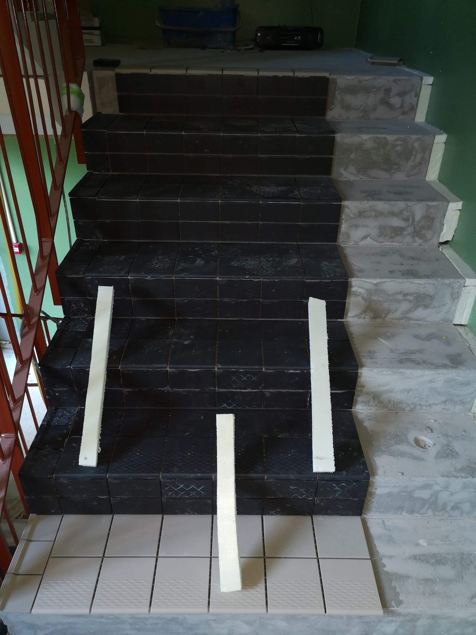 Schody w trakcie układania płytek: część stopni wykończona czarnymi płytkami, część w surowym betonie, białe listwy dystansowe między płytkami, na górze schodów radio.