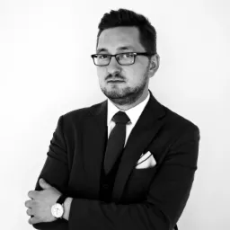 Portret mężczyzny w garniturze, okularach i z zegarkiem, z rękami skrzyżowanymi na piersiach, na białym tle, w czerni i bieli.