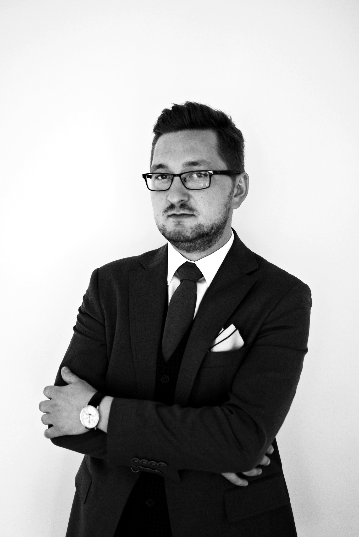 Portret mężczyzny w garniturze, okularach i z zegarkiem, z rękami skrzyżowanymi na piersiach, na białym tle, w czerni i bieli.