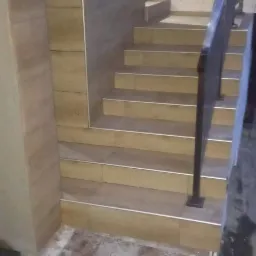 Schody wyłożone jasnymi płytkami ceramicznymi z metalowymi listwami krawędziowymi, z ciemną metalową barierką, ściany częściowo pokryte szarymi płytkami, widoczne ślady po fugowaniu na podłodze.