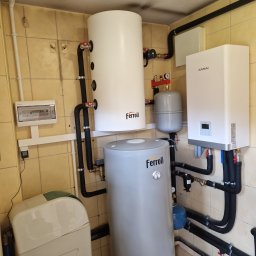 INSTALMATE Mateusz Ząbek - Dwa zbiorniki Ferroli różnej wielkości, biały wiszący piec Kaisai, instalacja hydrauliczna z czarnymi rurami, szary zbiornik wyrównawczy i zmiękczacz wody w pomieszczeniu z beżowymi płytkami.