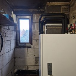 INSTALMATE Mateusz Ząbek - Biała pompa ciepła Kaisai z czarnymi rurami w pomieszczeniu technicznym z oknem i betonowymi ścianami.