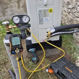 Podłączenie butli z azotem i manometru cyfrowego Testo 550s do zewnętrznej jednostki pompy ciepła Kaisai, widoczny także wkrętak akumulatorowy Makita, na tle ściany z kamienia.