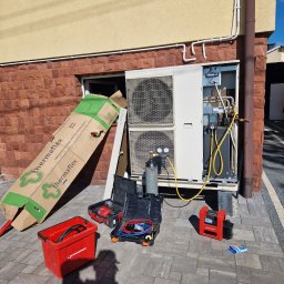 INSTALMATE Mateusz Ząbek - Montaż pompy ciepła przy budynku: jednostka zewnętrzna, butla z gazem, narzędzia Rothenberger. Widoczne okablowanie i rury. Częściowo otwarte okno piwniczne.