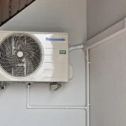 Zamontowany klimatyzator Panasonic na jasnej ścianie zewnętrznej, z widocznymi rurami odprowadzającymi i osłoną instalacji. Ujęcie z dołu, fragment dachu w tle.