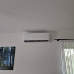 INSTALMATE Mateusz Ząbek - Biały klimatyzator ścienny w jasnym wnętrzu, z obrazem lasu i zasłoniętym oknem. Widoczny wyświetlacz z temperaturą. Minimalistyczny design, nowoczesne wykończenie.