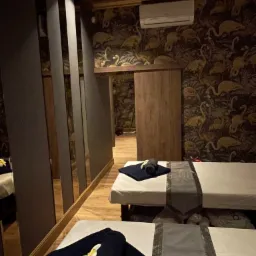 Przyciemnione wnętrze gabinetu masażu z dwoma przygotowanymi łóżkami, ciemne tapety w motywy tropikalne, klimatyzacja na ścianie, ciemne drewniane elementy wykończenia i dekoracyjne panele lustrzane.