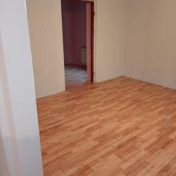 Krzysztof Barczycki - Pusty pokój po remoncie: świeżo położony parkiet w odcieniach brązu, jasnoszare ściany, widok na sąsiednie pomieszczenie przez otwarte drzwi.