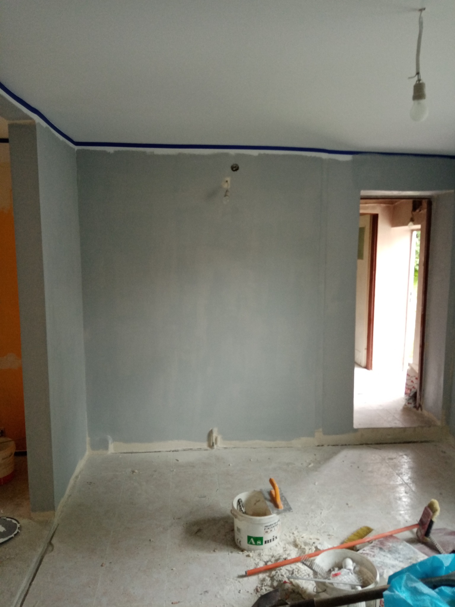 Świeżo pomalowane na szaro wnętrze pokoju z niebieską taśmą malarską przy suficie, widoczne narzędzia malarskie i wiadro z farbą na podłodze.