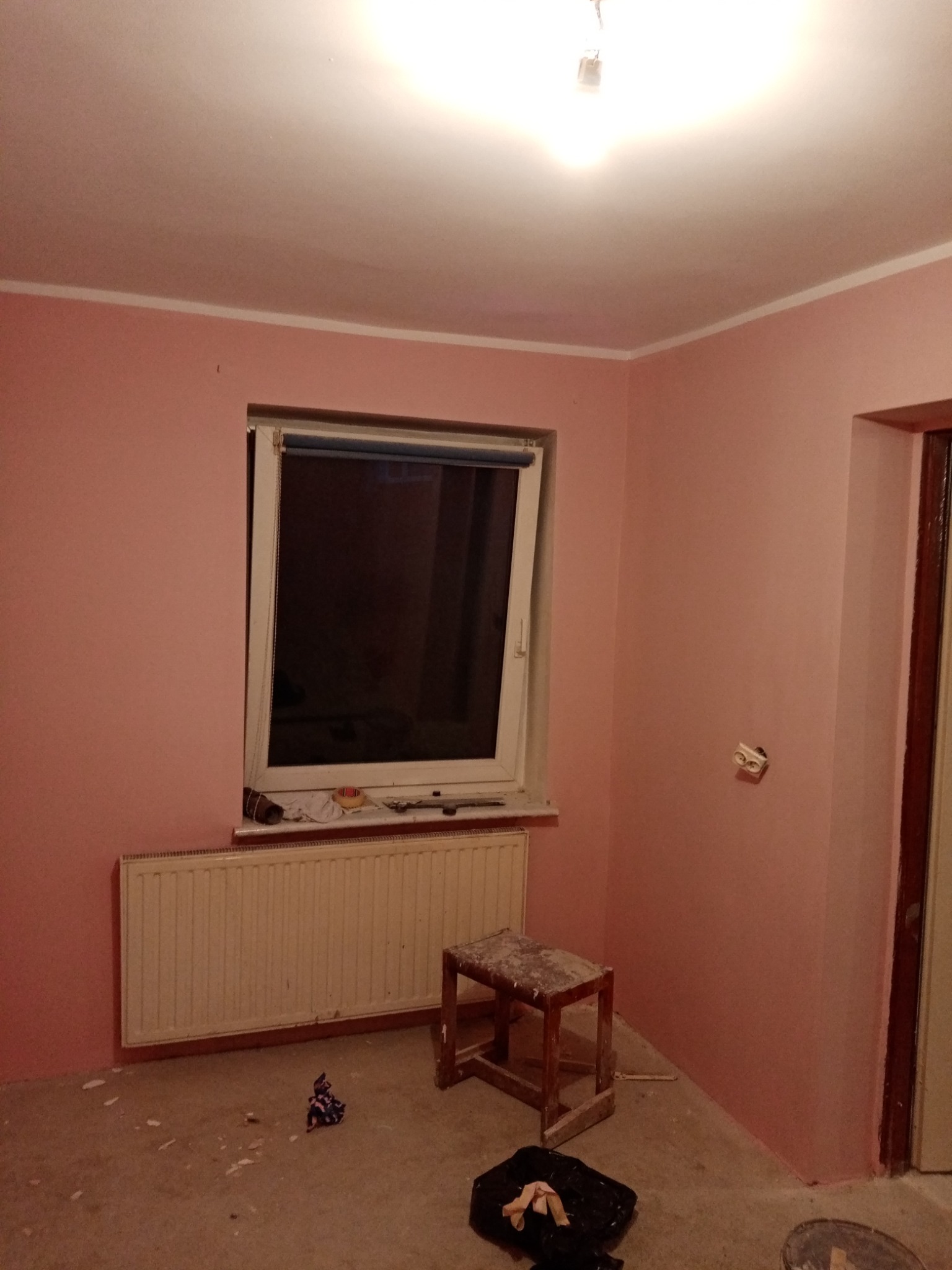 Pokój po świeżym pomalowaniu na różowo, widoczny grzejnik, okno z roletą, drewniany taboret i ślady farby na podłodze.
