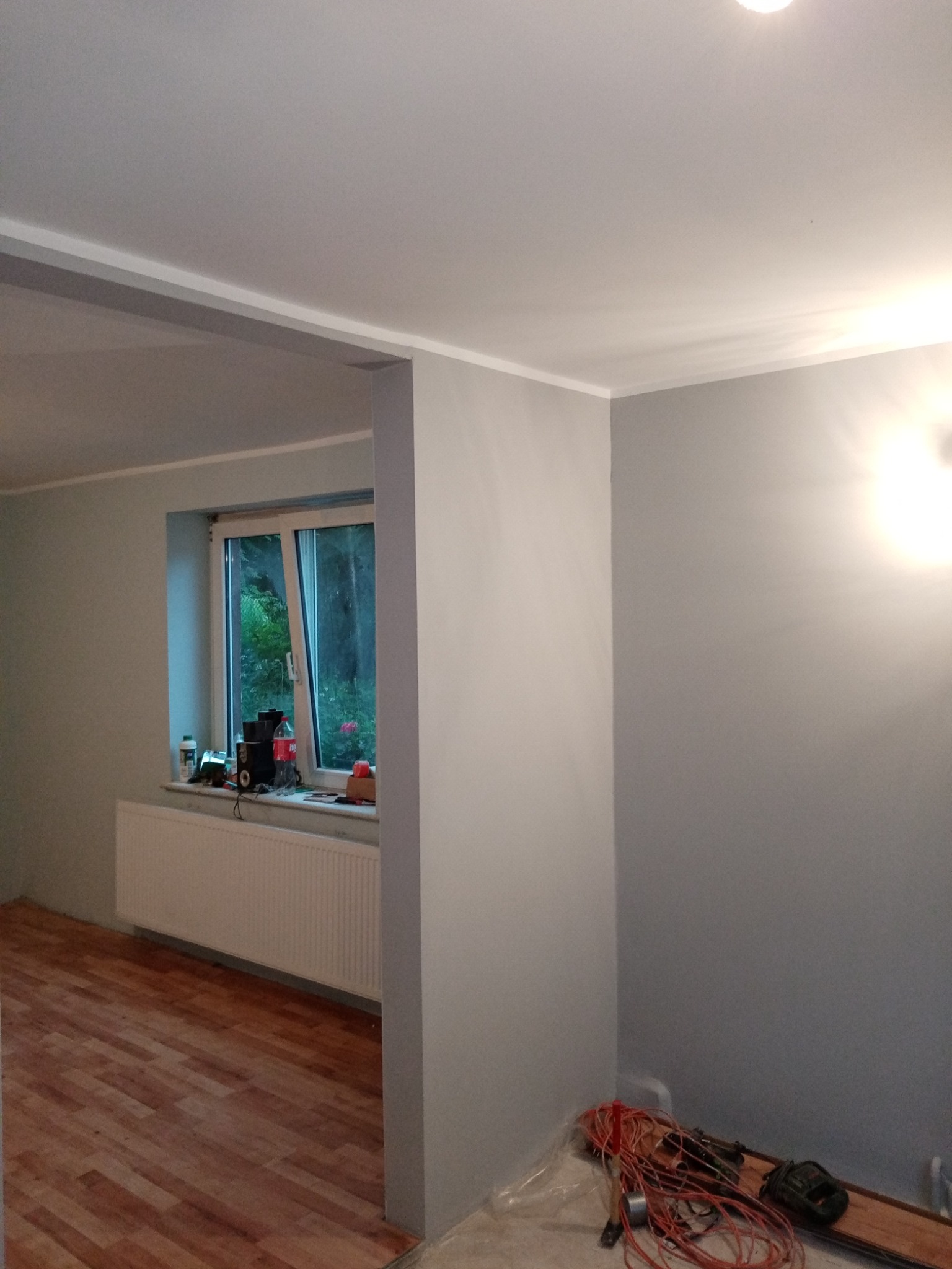 Świeżo pomalowane na szaro wnętrze pokoju z drewnianą podłogą i grzejnikiem pod oknem, widoczne narzędzia i przewody elektryczne na podłodze, przygotowane do dalszych prac wykończeniowych.