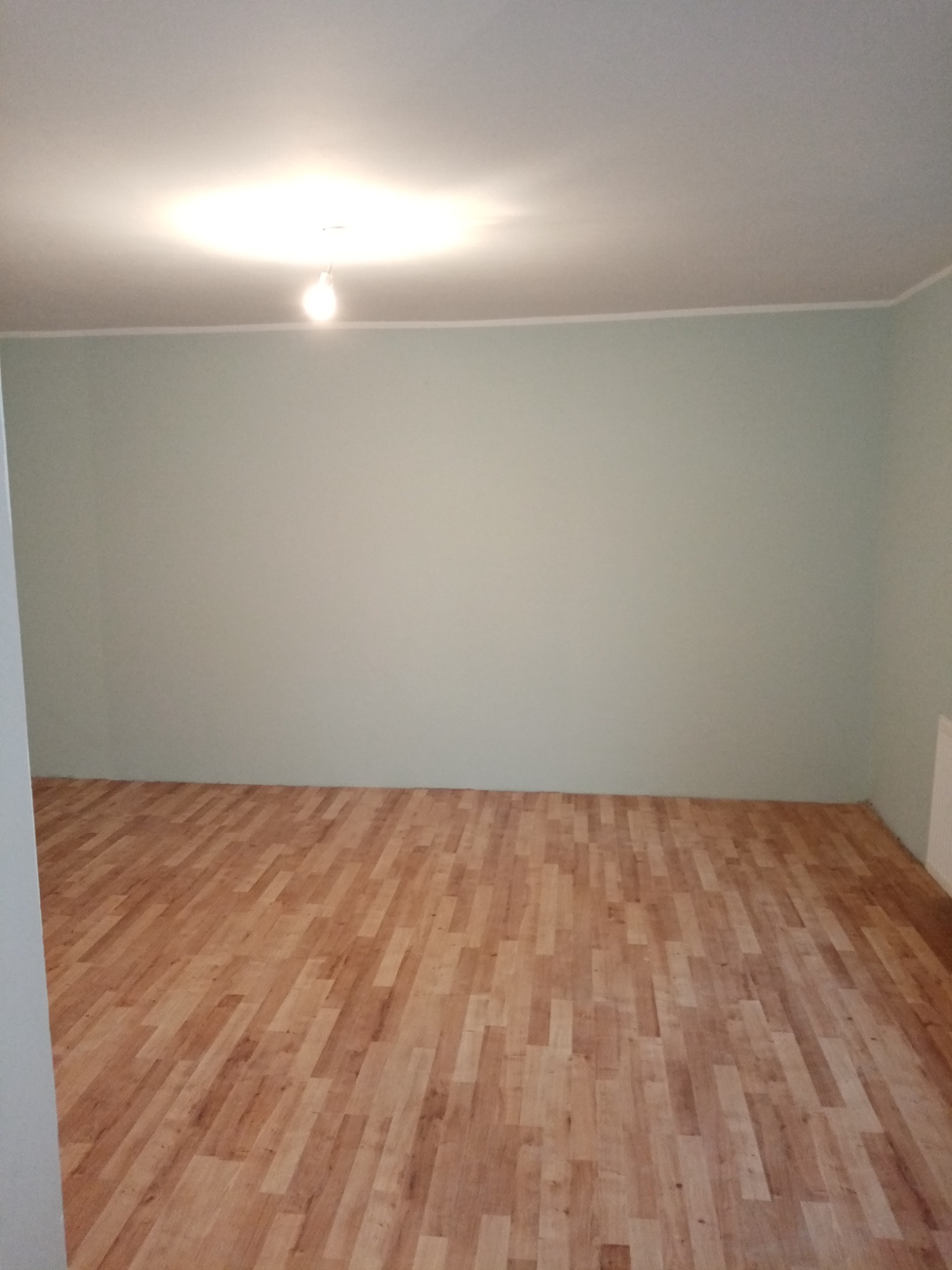 Pusty pokój z drewnianą podłogą ułożoną w jodełkę i świeżo pomalowanymi ścianami na jasnozielony kolor, widoczna żarówka na suficie.