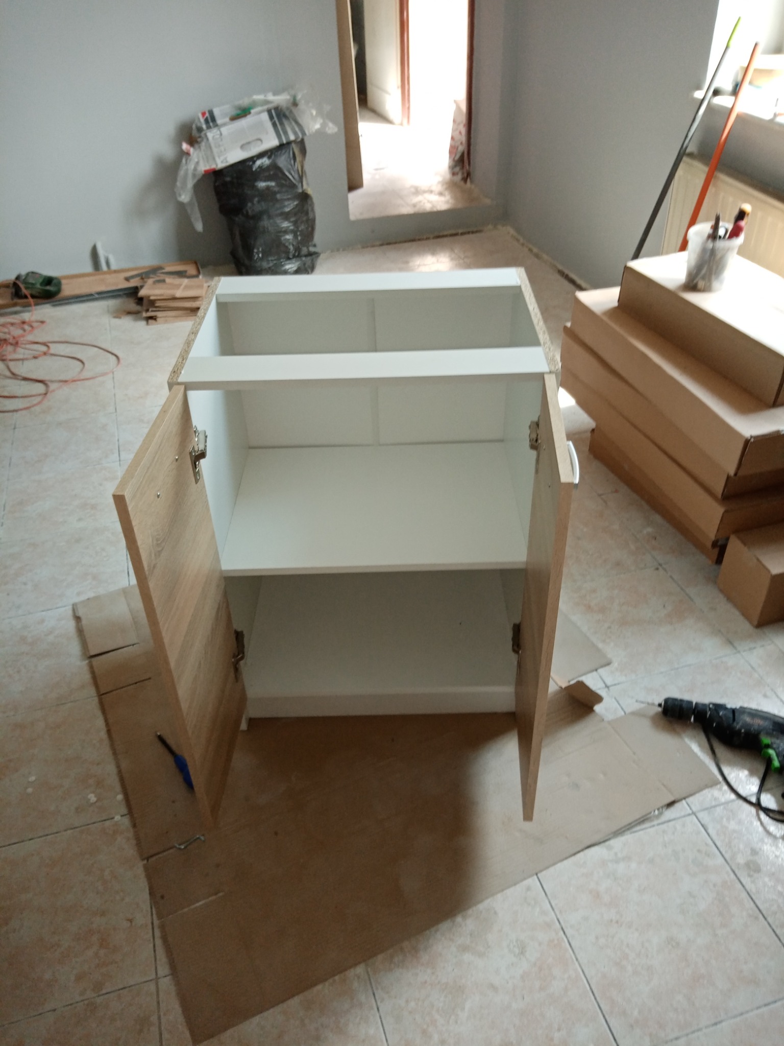 Szafka z otwartymi drzwiczkami w trakcie montażu, widoczne kartony, wiertarka, śmieci i narzędzia na podłodze z płytek.