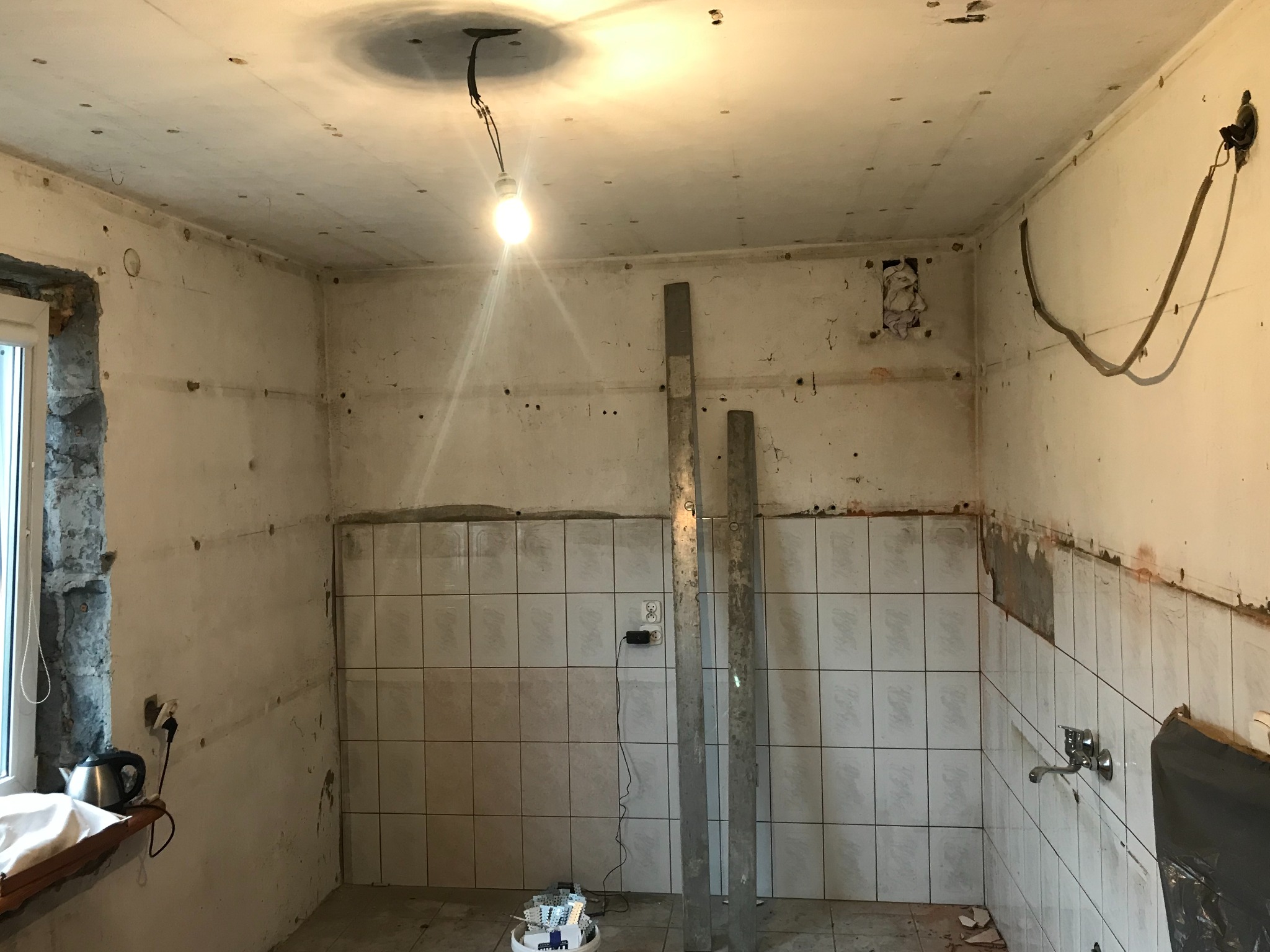 Wnętrze kuchni w trakcie prac przygotowawczych do remontu: widoczne ściany z częściowo usuniętymi płytkami, odsłonięte przewody elektryczne, surowy sufit z wiszącą żarówką, metalowe profile...