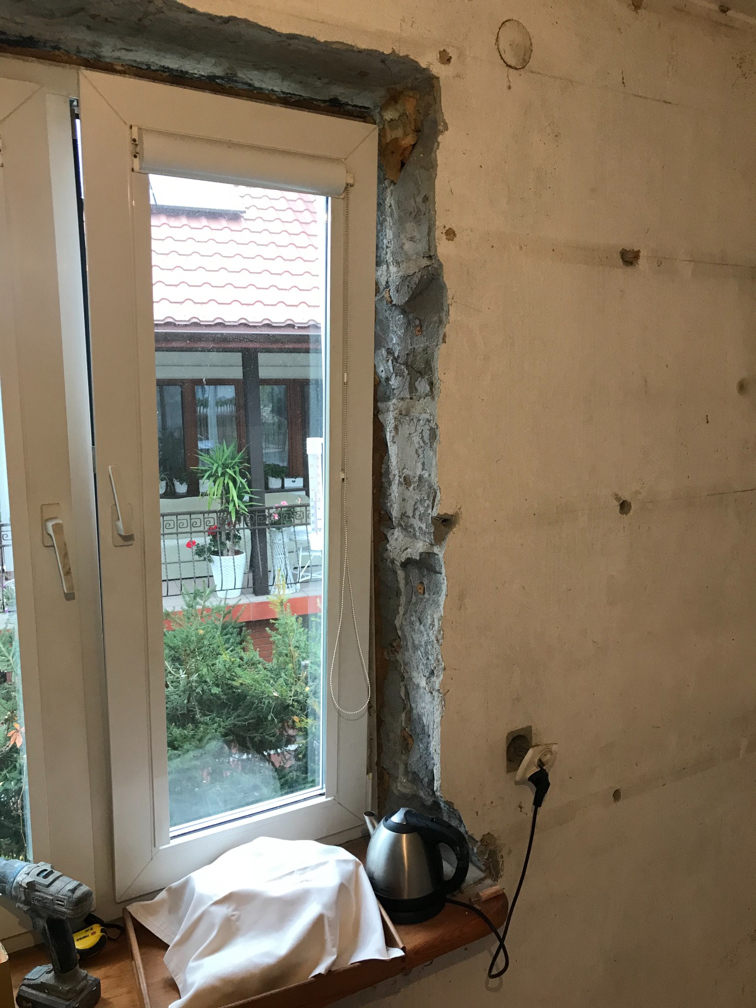 Przygotowanie do prac remontowych: odsłonięty mur przy oknie, widoczny kabel zasilający, czajnik elektryczny i wiertarka na parapecie.