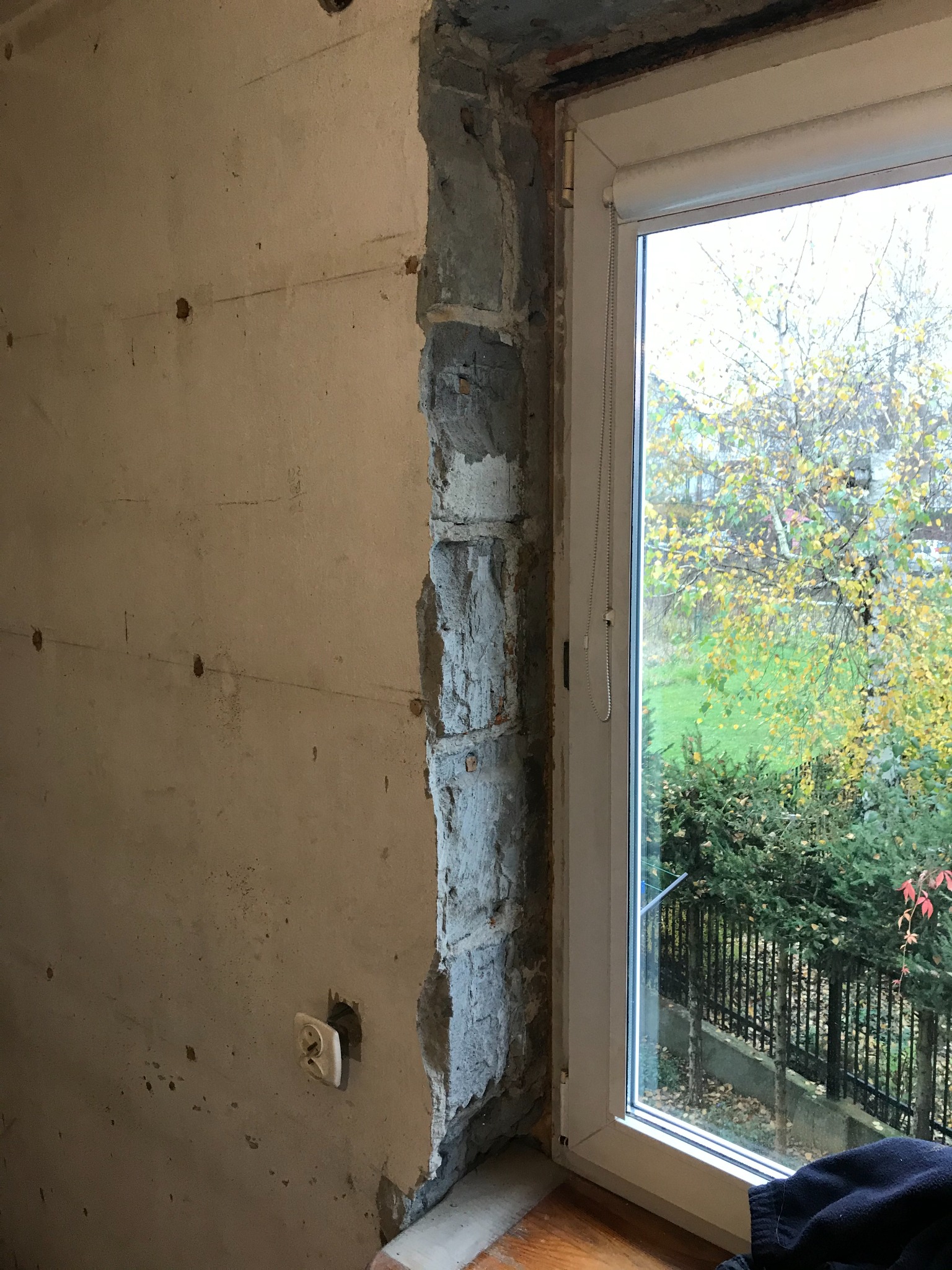 Przygotowanie otworu okiennego do osadzenia ościeżnicy: surowa ściana z widocznymi śladami po kuciu, odsłonięta cegła i wystający kabel elektryczny obok gniazdka, widok na jesienne drzewa za oknem.