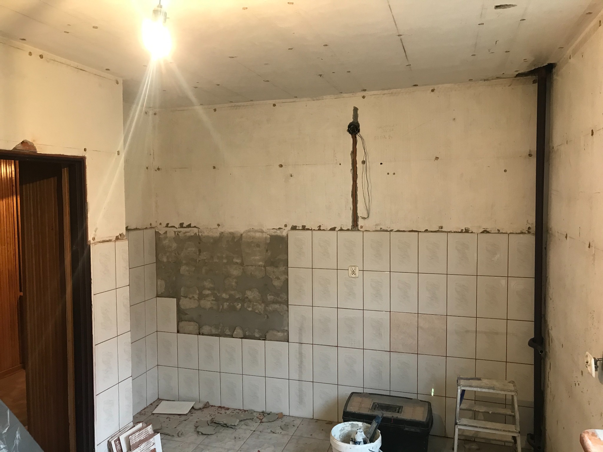 Wnętrze w trakcie prac przygotowawczych do położenia płytek, widoczne fragmenty ściany z nałożonym klejem, częściowo ułożone płytki, narzędzia i drabina.
