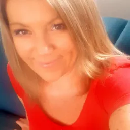 Portret uśmiechniętej kobiety z blond włosami w czerwonej bluzce, siedzącej na niebieskiej sofie.