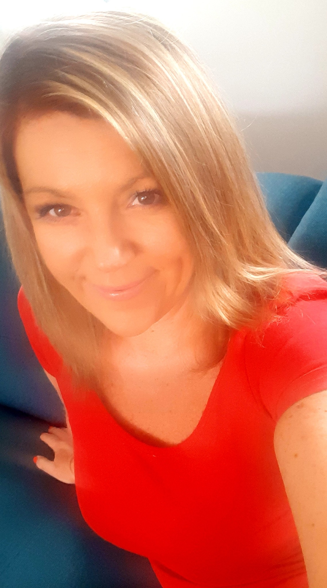 Portret uśmiechniętej kobiety z blond włosami w czerwonej bluzce, siedzącej na niebieskiej sofie.