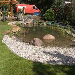 Krzysztof Bogucki usługi ogrodnicze - Budowa oczka wodnego z wykończeniem z białych kamieni i trawy z rolki, w tle pracownicy i czerwony samochód dostawczy.