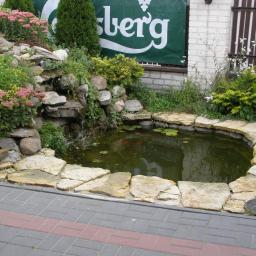 Krzysztof Bogucki usługi ogrodnicze - Oczko wodne z kaskadą wodną obłożone kamieniami, otoczone roślinnością ozdobną i wykończone brukowaną ścieżką, w tle widoczny baner reklamowy.