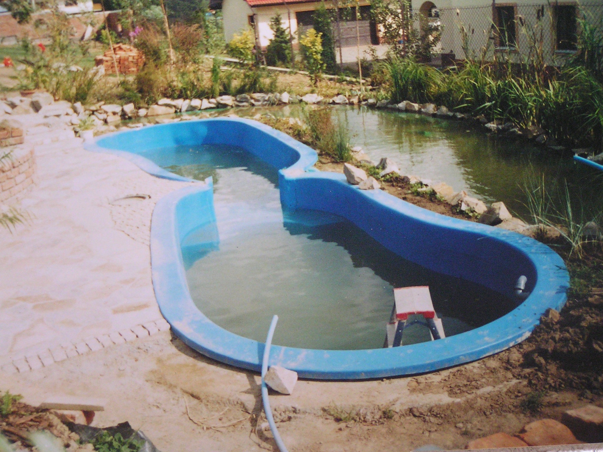 Formowany, niebieski zbiornik wodny w trakcie instalacji, otoczony kamieniami i roślinnością, z widocznym fragmentem trawnika i zabudowań w tle. Woda w zbiorniku lekko mętna, z boku leży biały...