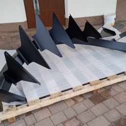 Allmet Magdalena Weymann-Wypchlak - Elementy metalowe w kolorze czarnym, prawdopodobnie części ogrodzenia lub balustrady, ułożone na palecie wyłożonej folią bąbelkową przed budynkiem z brązowymi drzwiami.