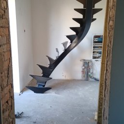 Allmet Magdalena Weymann-Wypchlak - Czarne, metalowe schody spiralne z trójkątnymi stopniami, zamontowane w surowym wnętrzu z widoczną instalacją elektryczną i wodną, widziane zza futryny z odsłoniętą cegłą.