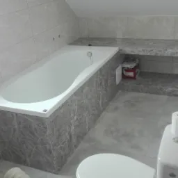 Ukończona łazienka na poddaszu ze skośnym sufitem, wykończona płytkami imitującymi marmur, z wanną zabudowaną w płytkach, toaletą i grzejnikiem olejowym.