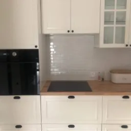 Biała zabudowa kuchenna z piekarnikiem w zabudowie, drewnianym blatem i szarymi płytkami na ścianie, uzupełniona o pojemnik na chleb.