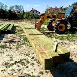 Żółty wózek widłowy podnosi długie, zielone, kwadratowe belki drewniane na placu budowy w słoneczny dzień.