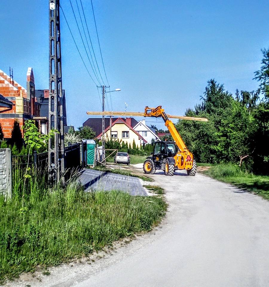 Żółta ładowarka teleskopowa transportuje drewniane belki na tle budowy domu z czerwonej cegły i zieleni.