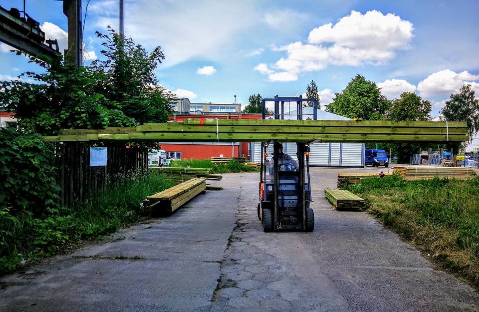 Wózek widłowy transportuje pakiet impregnowanej tarcicy na terenie składu drewna, widoczne składowane deski, budynki w tle i błękitne niebo z chmurami.