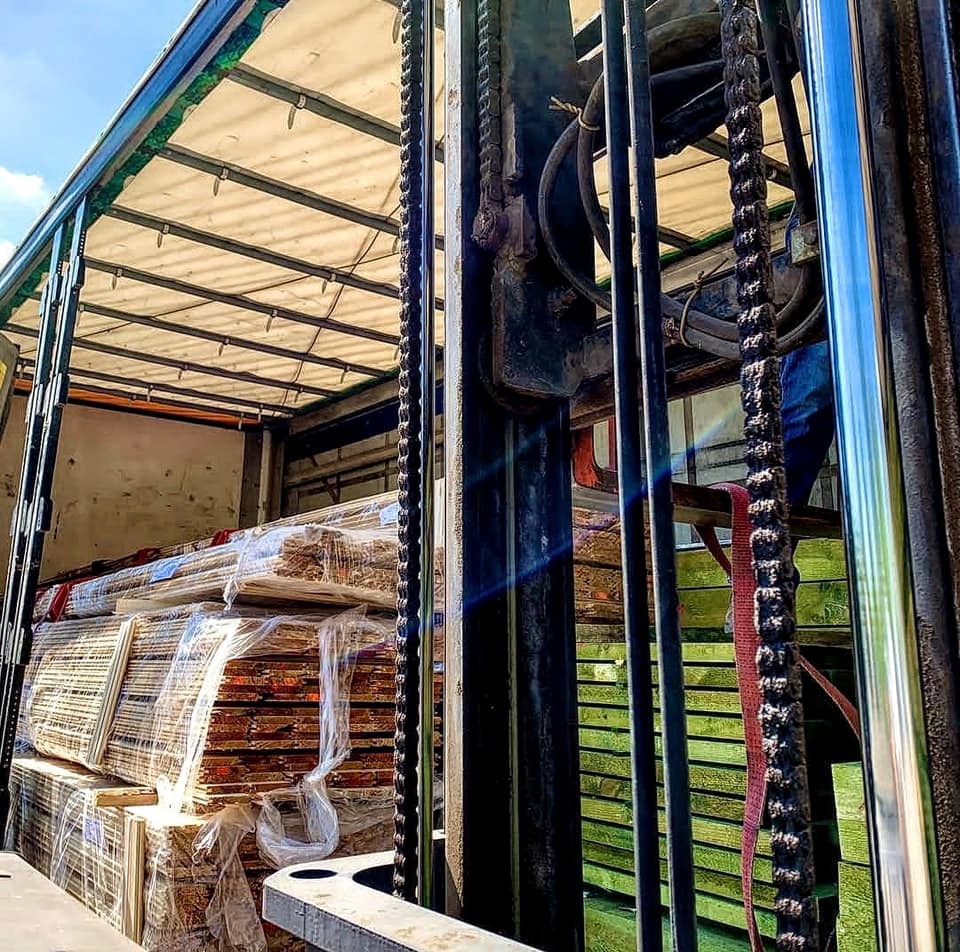 Wózek widłowy załadowuje pakiet tarcicy owinięty folią do wnętrza naczepy ciężarówki, widoczne łańcuchy wózka i elementy konstrukcyjne naczepy.