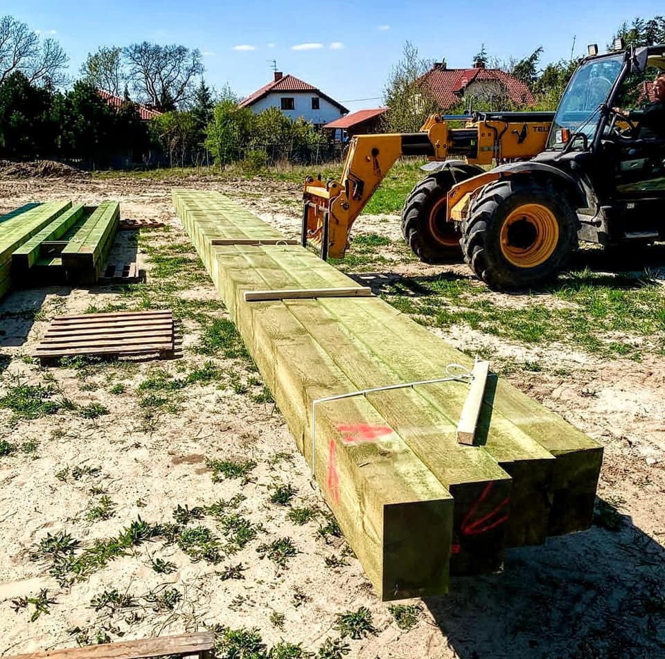 Żółty wózek widłowy podnosi długie, zielone, kwadratowe belki drewniane na placu budowy w słoneczny dzień.