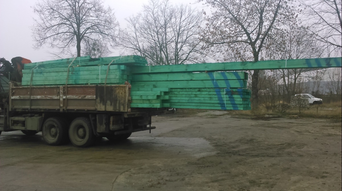 Ciężarówka z dźwigiem załadowana zieloną, zabezpieczoną tarcicą z niebieskimi oznaczeniami, na tle drzew i pochmurnego nieba w Poznaniu.