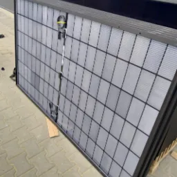 Stos paneli fotowoltaicznych z czarną ramą, zabezpieczonych taśmą, leżących na kostce brukowej. Widoczne złącza. Przygotowane do montażu.