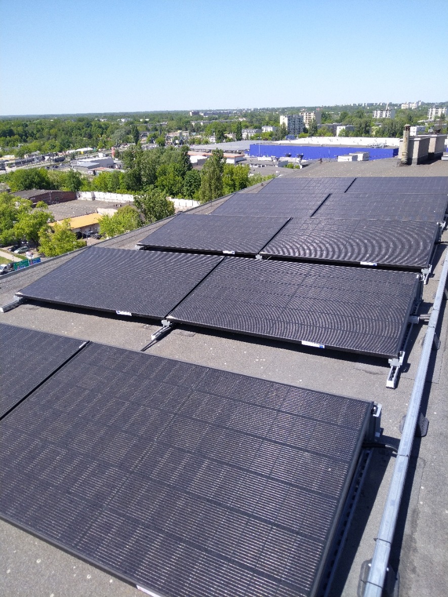 Rząd paneli słonecznych na płaskim dachu budynku przemysłowego, z widokiem na zieleń miejską i błękitne niebo w tle.