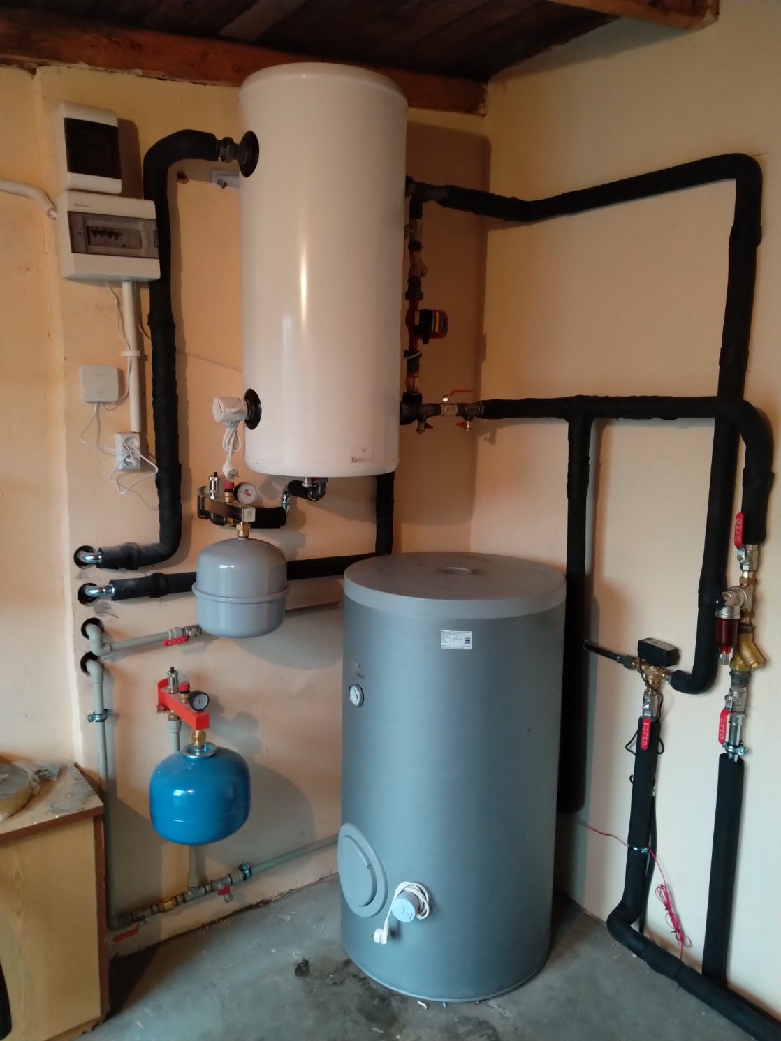 Instalacja centralnego ogrzewania z dwoma zbiornikami: białym wiszącym i szarym stojącym, połączone czarnymi rurami w izolacji. Widoczny także mały niebieski zbiornik wyrównawczy i rozdzielnia...