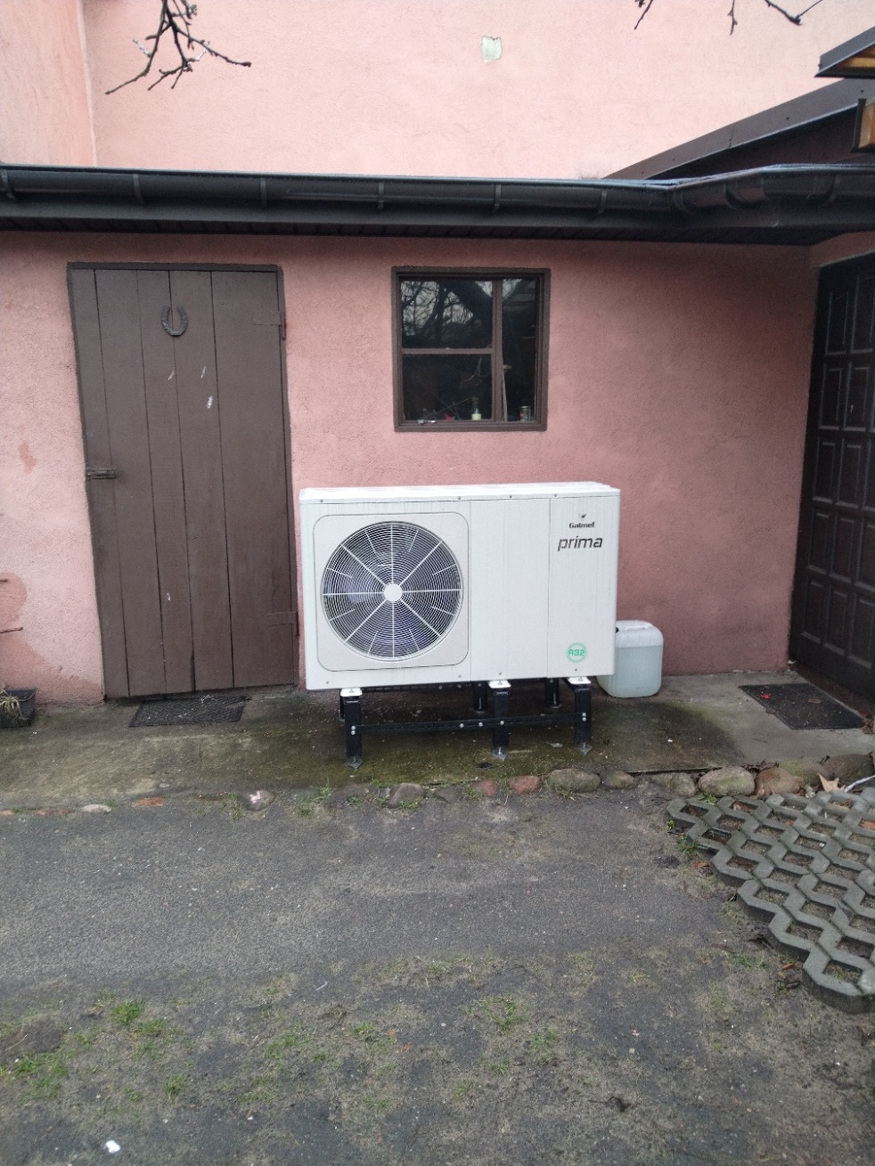 Biała pompa ciepła Galmet Prima zamontowana na czarnym stelażu przed budynkiem z różową elewacją, widoczne drzwi z podkową i okno.