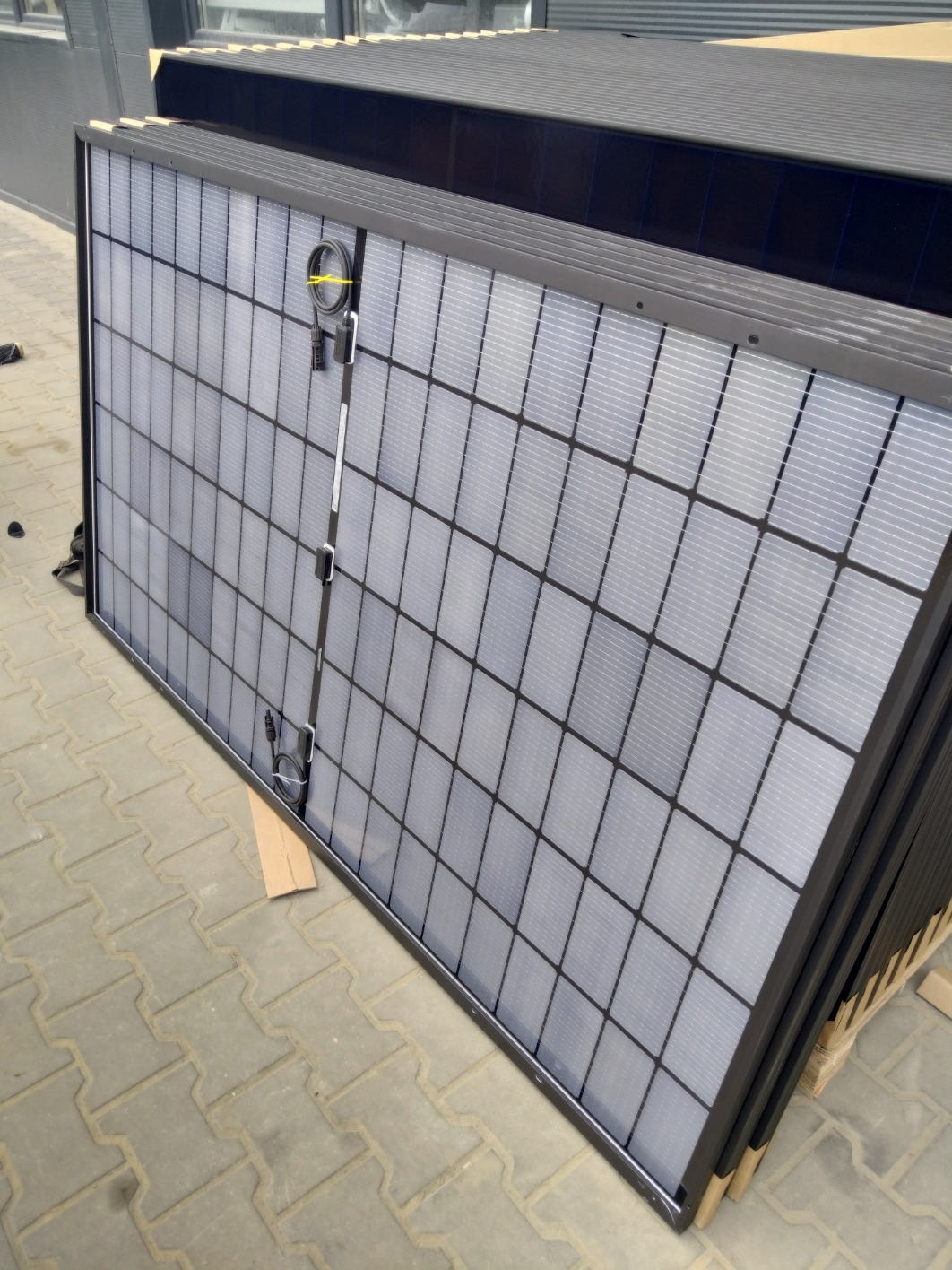 Stos paneli fotowoltaicznych z czarną ramą, zabezpieczonych taśmą, leżących na kostce brukowej. Widoczne złącza. Przygotowane do montażu.