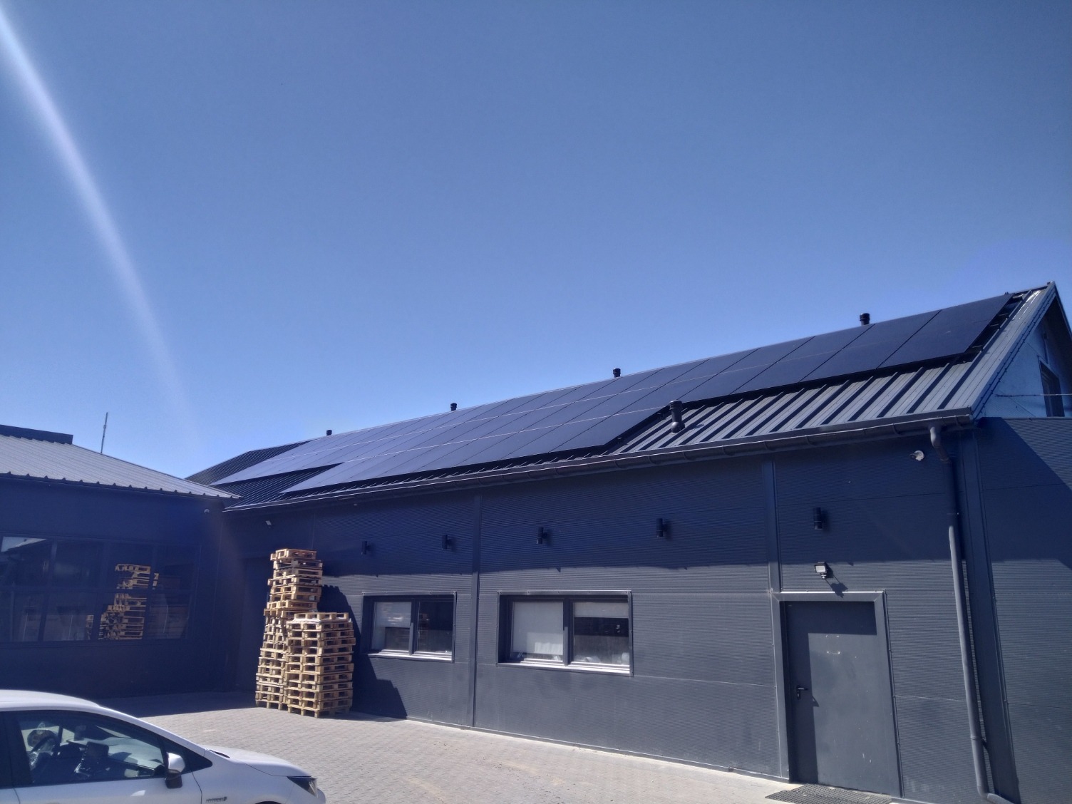 Ciemnoszare panele fotowoltaiczne na dachu budynku przemysłowego w słoneczny dzień. Na pierwszym planie stos palet i fragment białego samochodu.