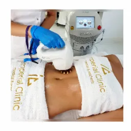 Pacjentka leżąca na stole zabiegowym, poddawana zabiegowi modelowania sylwetki przy użyciu urządzenia z głowicą. Widoczne rękawice ochronne osoby wykonującej zabieg oraz logo kliniki na ręczniku.