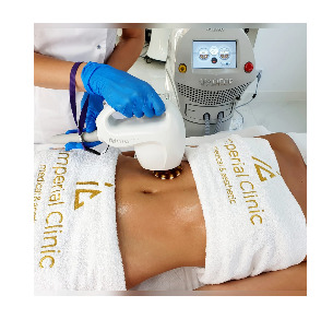 Pacjentka leżąca na stole zabiegowym, poddawana zabiegowi modelowania sylwetki przy użyciu urządzenia z głowicą. Widoczne rękawice ochronne osoby wykonującej zabieg oraz logo kliniki na ręczniku.