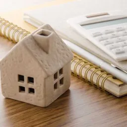 Ceramiczny model domu stoi obok kalkulatora i notatnika ze złotą spiralą na drewnianym biurku.