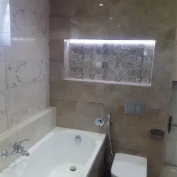 Wykończona łazienka w beżowych płytkach z dekoracyjną wnęką nad wanną, podświetloną listwą LED, wisząca toaleta i chromowana bateria wannowa.