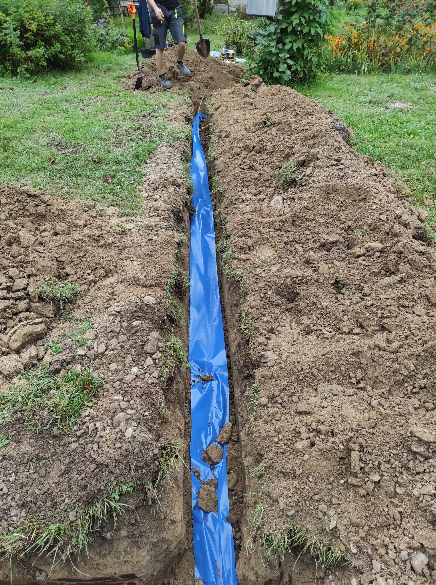 Wykopany rów w ziemi wyłożony niebieską folią, widoczne narzędzia i osoba pracująca w oddali, przygotowanie pod instalację.