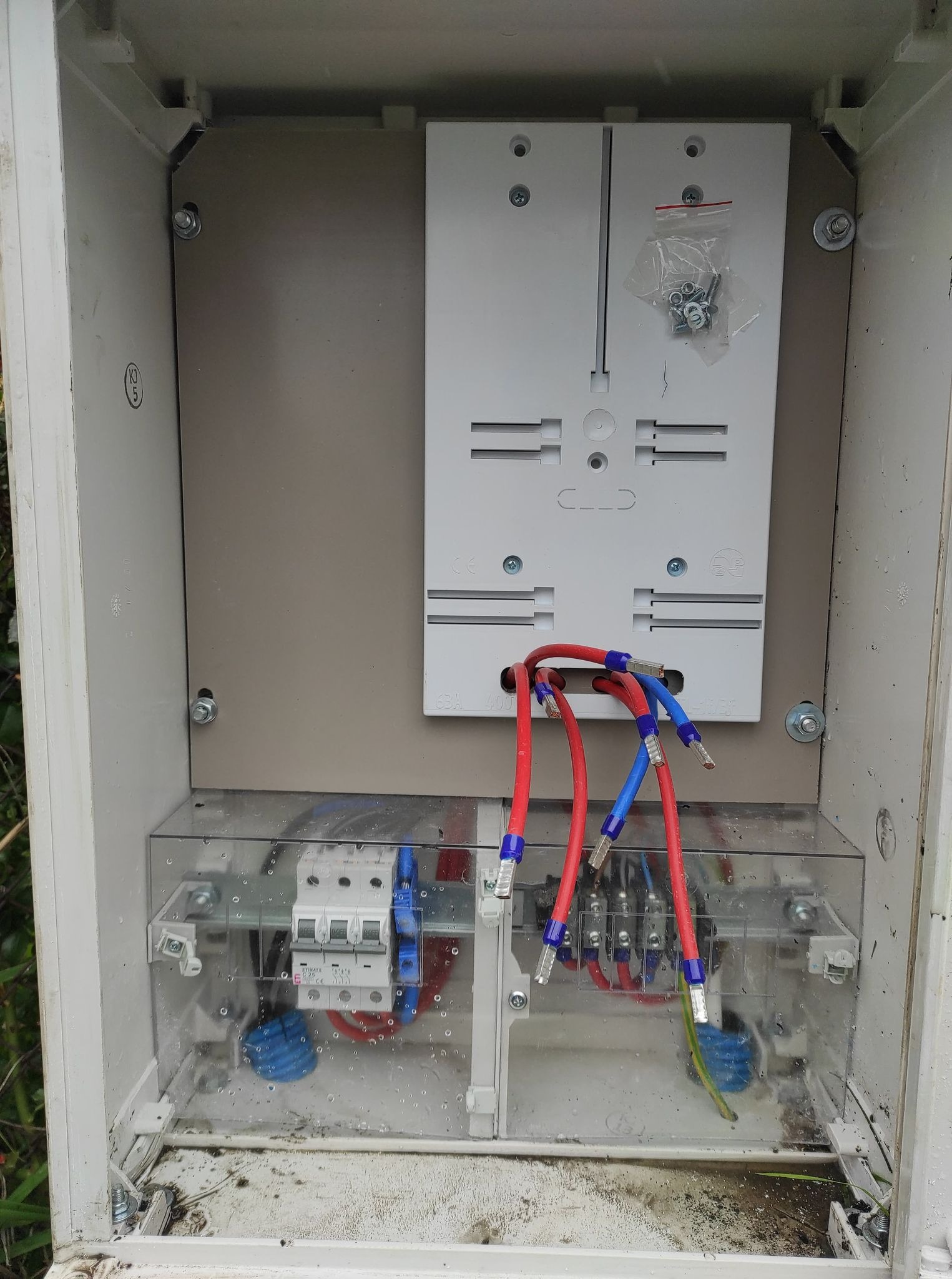 Wnętrze skrzynki elektrycznej z zamontowanymi bezpiecznikami i przewodami zakończonymi tulejkami, widoczne ślady wilgoci w dolnej części obudowy.